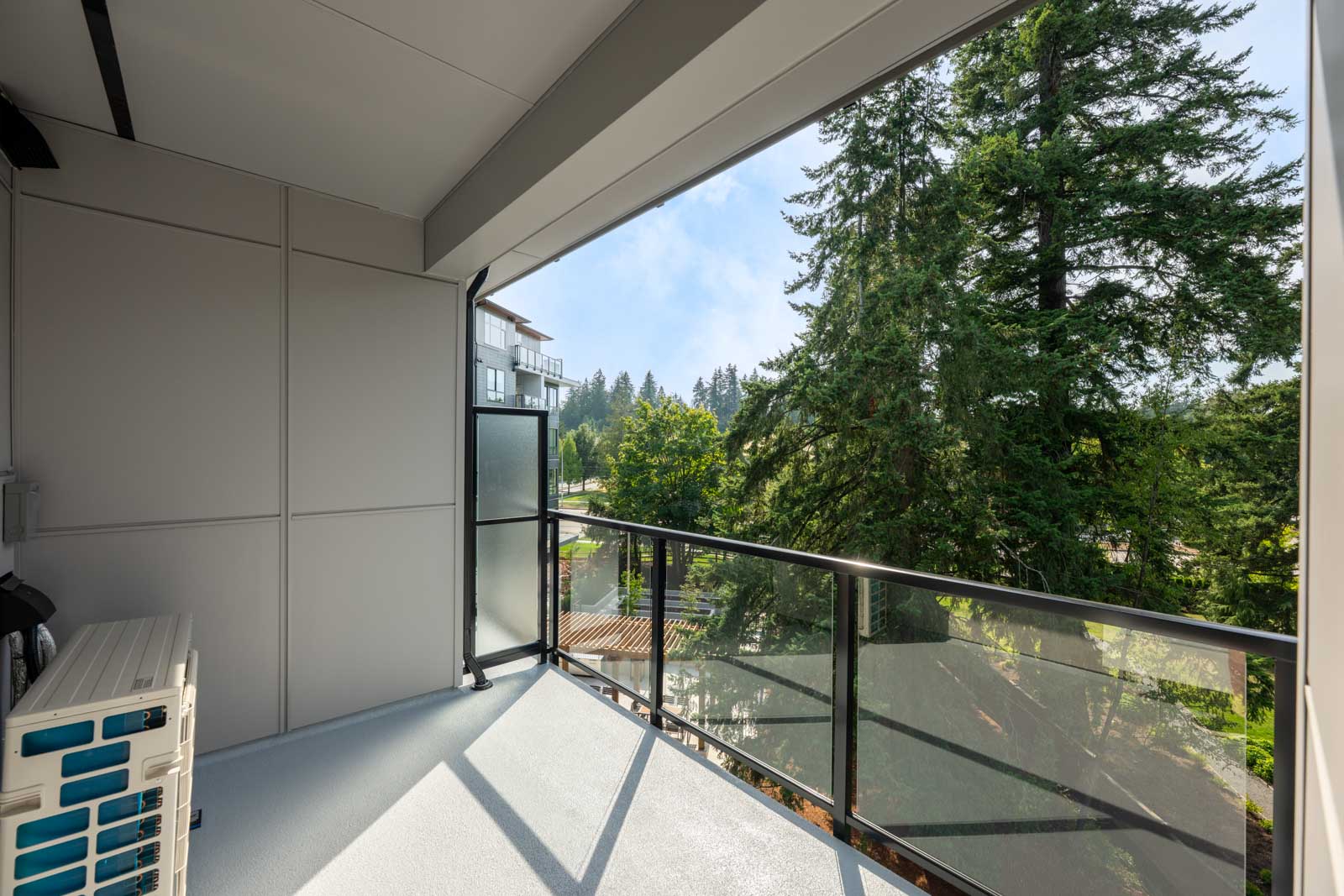 A modern balcony with glass railing overlooks tall green trees and a nearby building, under a clear blue sky.