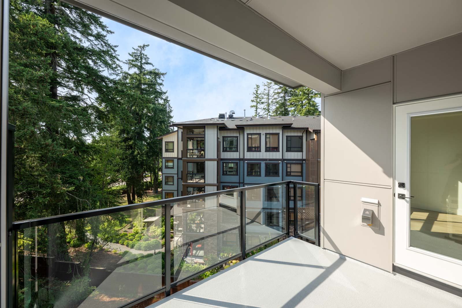 View from a modern balcony with glass railing, overlooking a landscaped courtyard and a multi-story apartment building surrounded by tall trees.