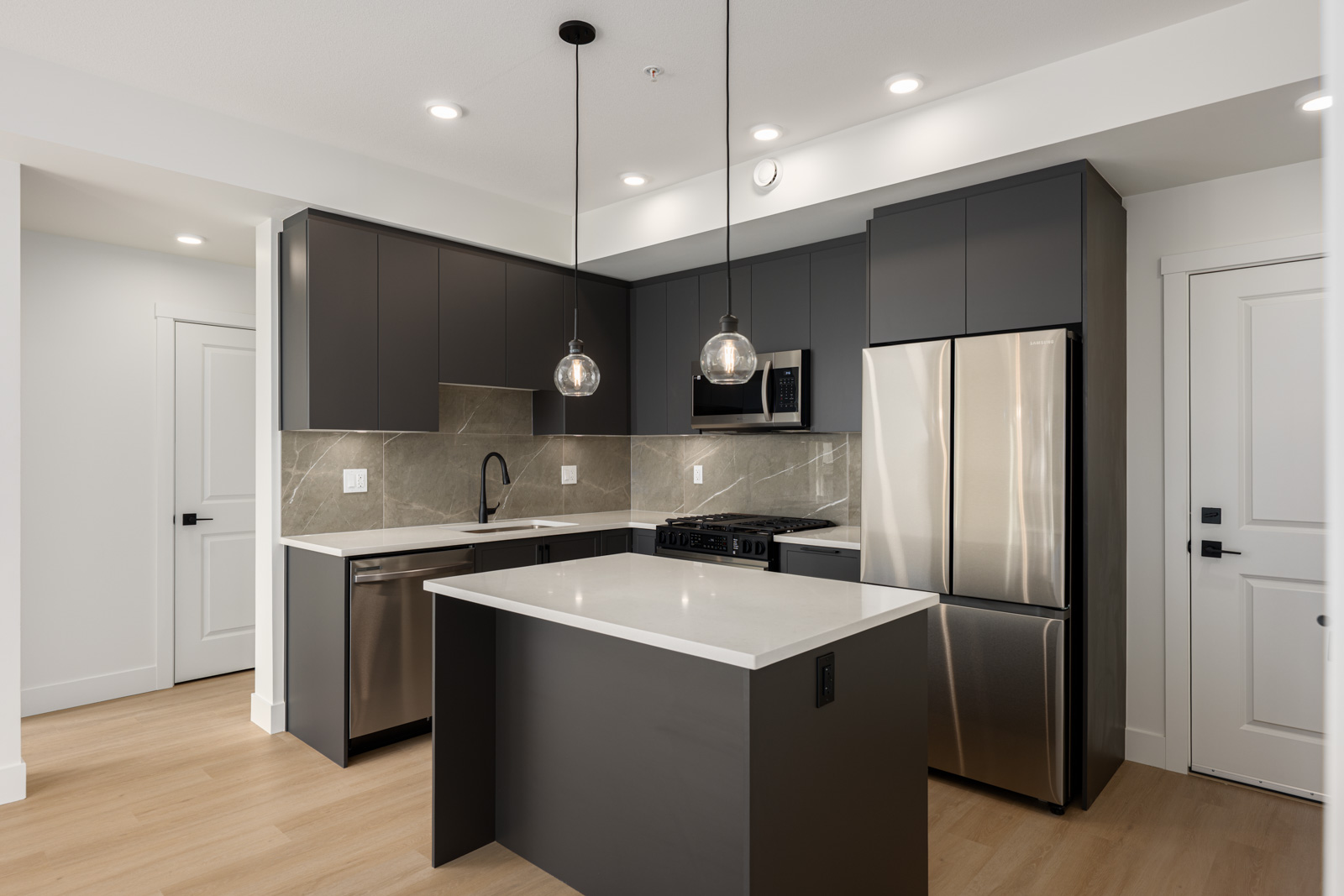 Modern kitchen with dark cabinets, stainless steel appliances, quartz countertops, tiled backsplash, light wood flooring, and two pendant lights over a central island.