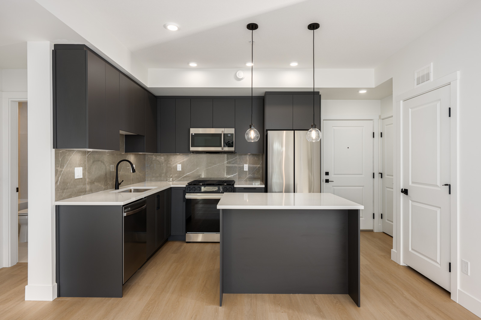 Modern kitchen with dark cabinets, stainless steel appliances, white countertops, pendant lights, and light wood flooring. Island is centered with two hanging lights above.