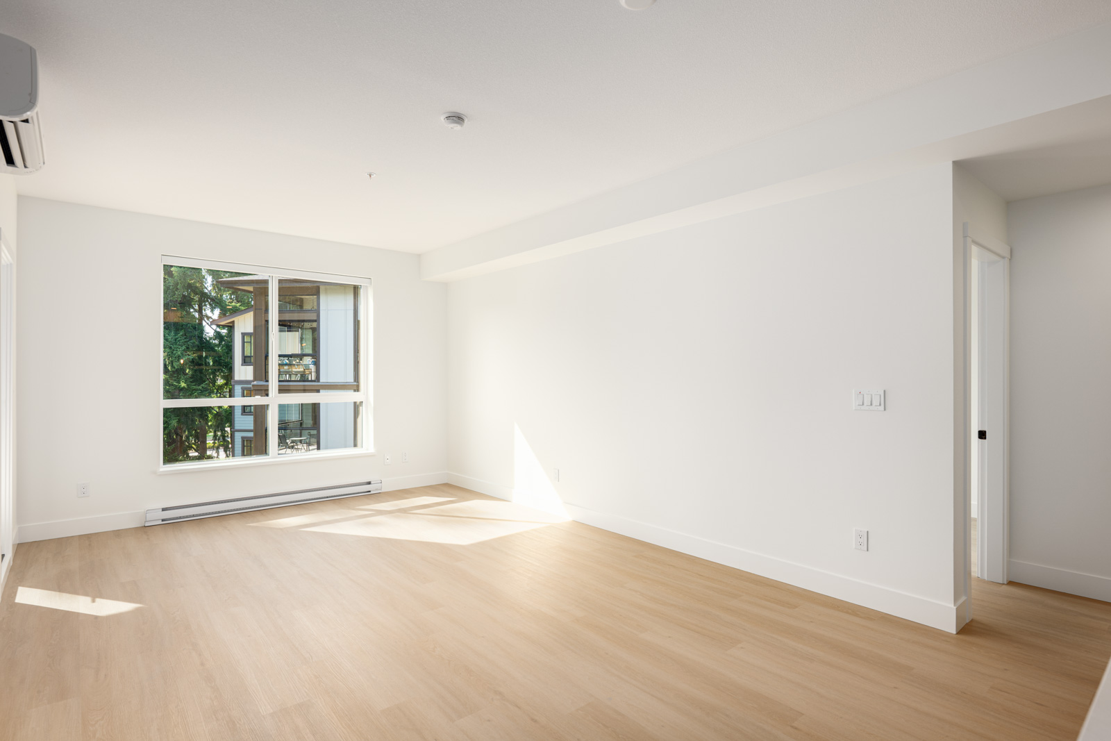 An empty room with light wood flooring, white walls, a large window, and a door leading to another space; natural light fills the room.