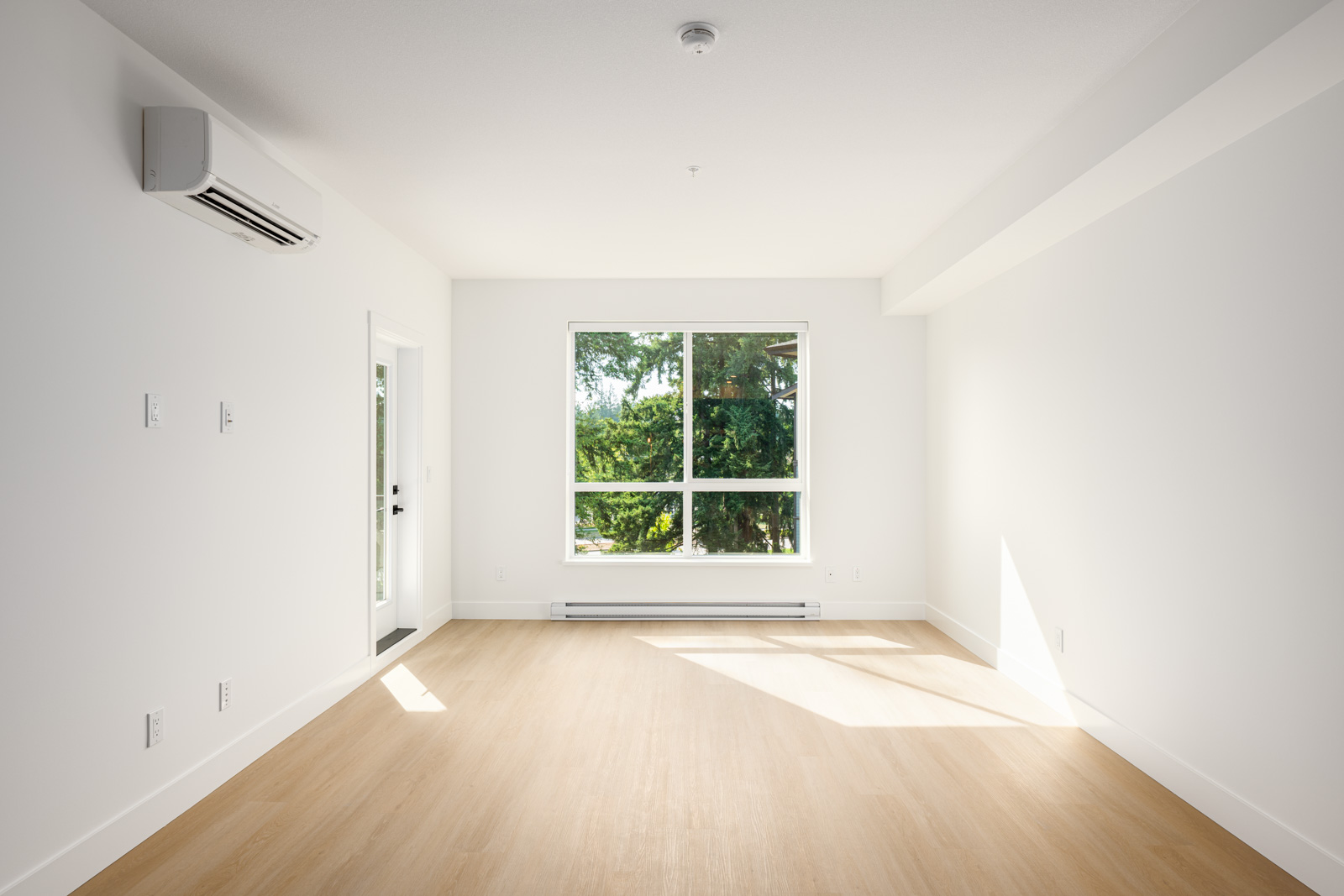 A bright, empty room with white walls, light wood flooring, a large window, a door to the outside, and an air conditioning unit mounted on the wall.