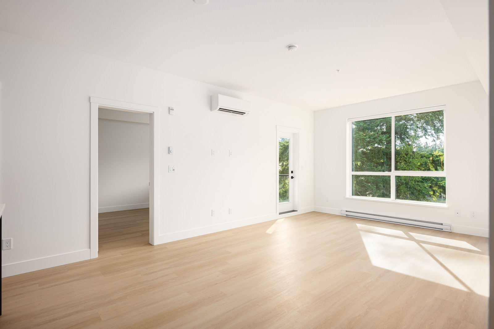 Bright, empty room with light wood floors, white walls, a large window with green trees outside, an air conditioning unit, and a door leading to another room.