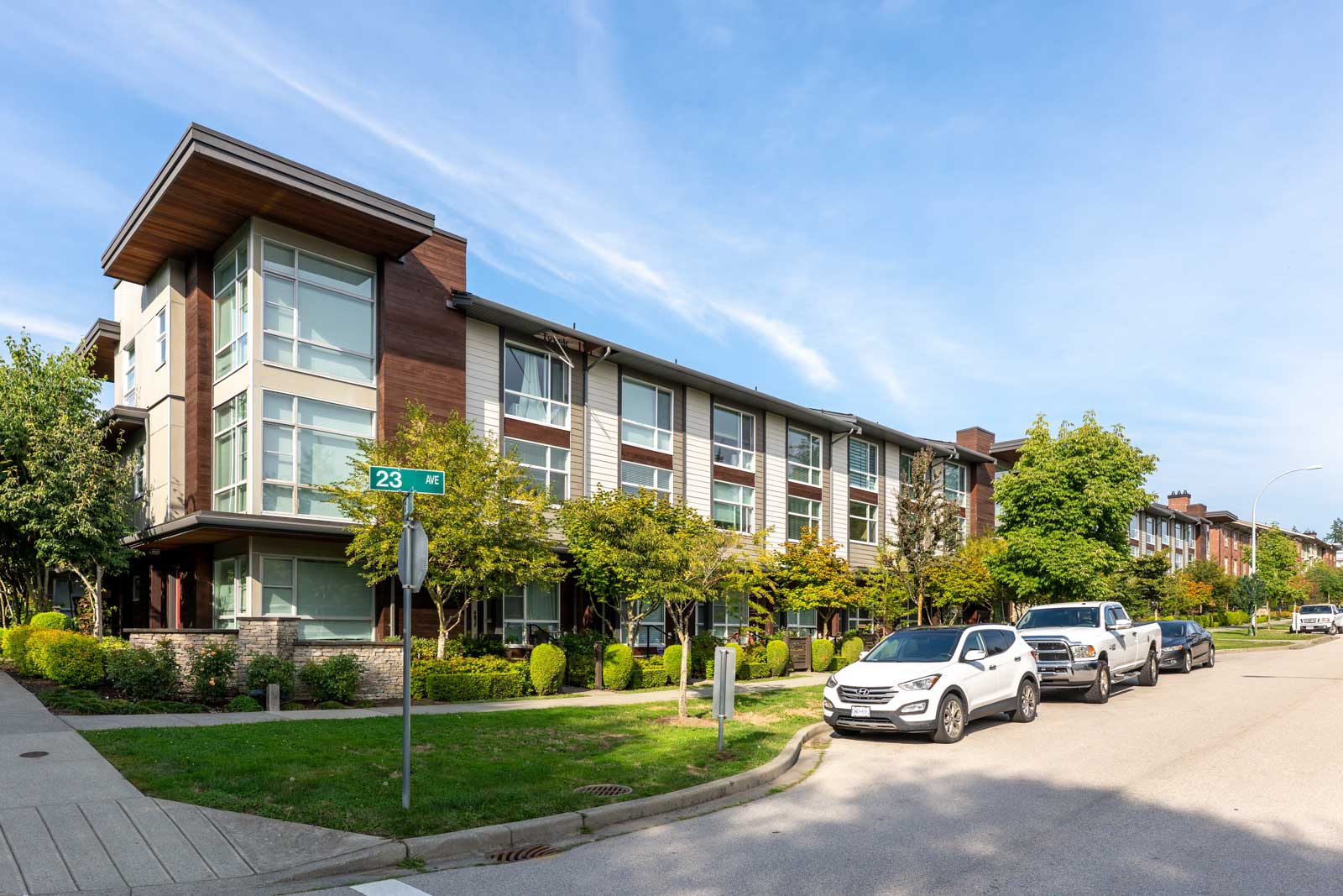Three vehicles are parked on a residential street beside a modern three-story apartment building with large windows and trees lining the sidewalk.