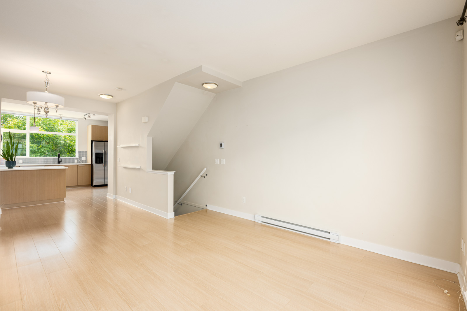 Unfurnished living room with light wood flooring, neutral walls, and a staircase leading down. Kitchen with stainless steel appliances and large window visible in the background.