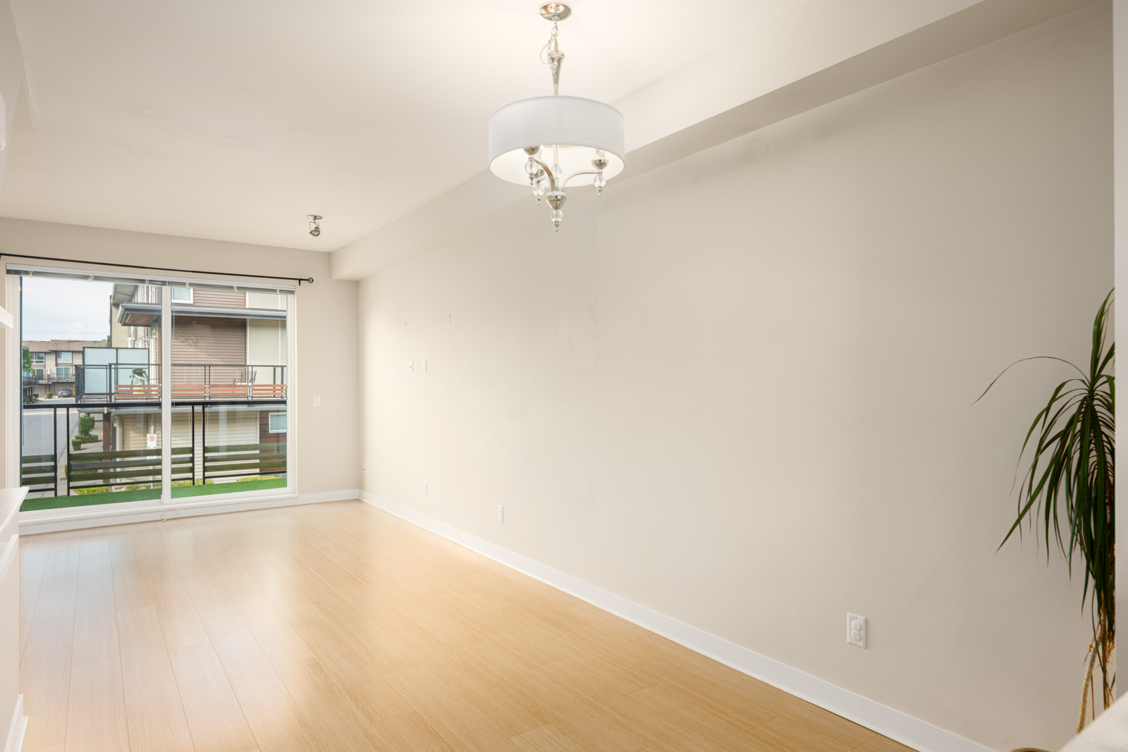 An empty room with light wood flooring, white walls, a modern chandelier, a large window with sliding glass doors, and a potted plant in the corner.