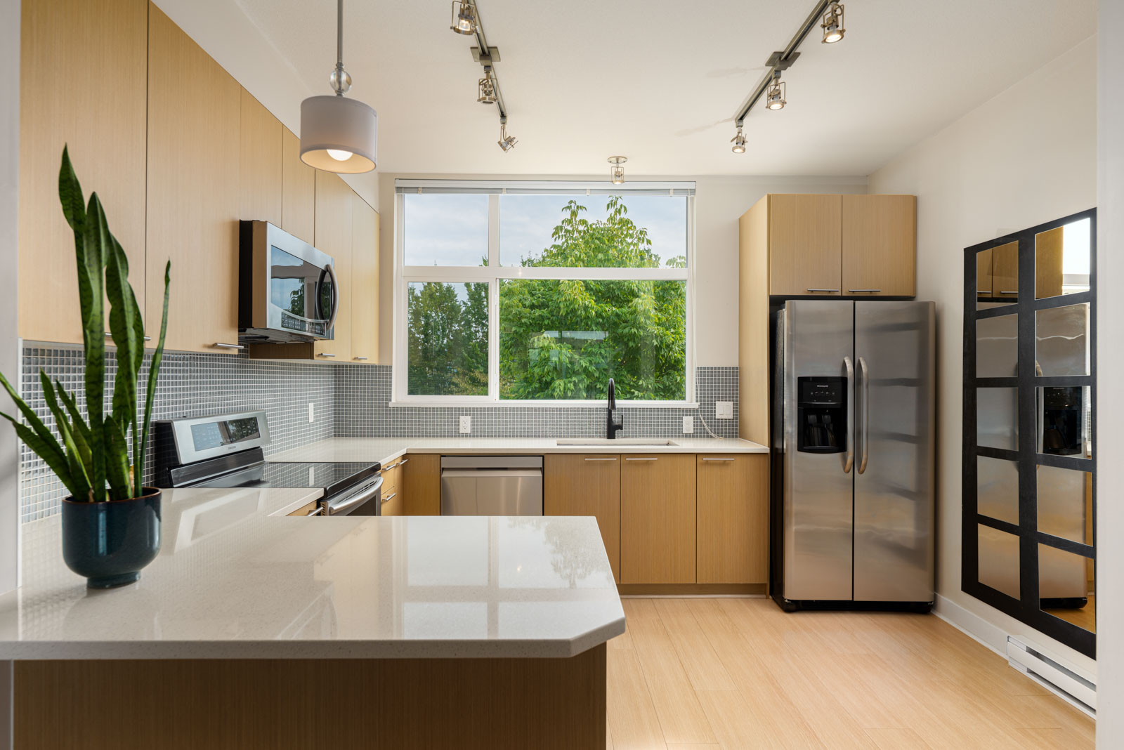 Modern kitchen with light wood cabinets, stainless steel appliances, a large window, and a potted plant on a white island countertop.