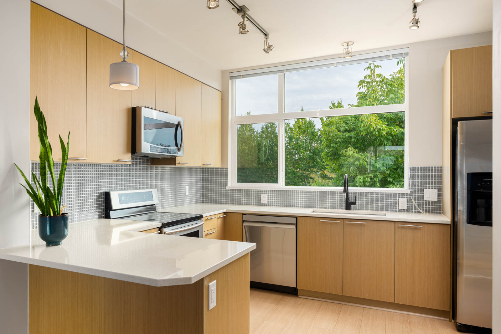 Modern kitchen with light wood cabinets, stainless steel appliances, a potted plant on the counter, mosaic tile backsplash, and large window with trees visible outside.