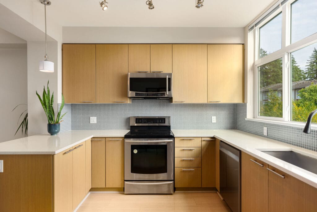 Modern kitchen with light wood cabinets, stainless steel appliances, mosaic tile backsplash, large window, and a potted plant on the counter.
