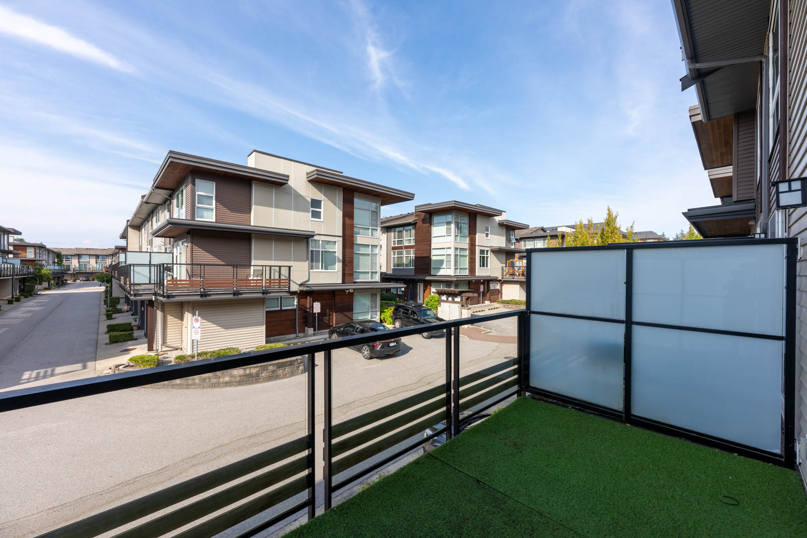 View from a balcony with artificial grass flooring, overlooking a residential street lined with modern multi-story townhouses under a clear blue sky.