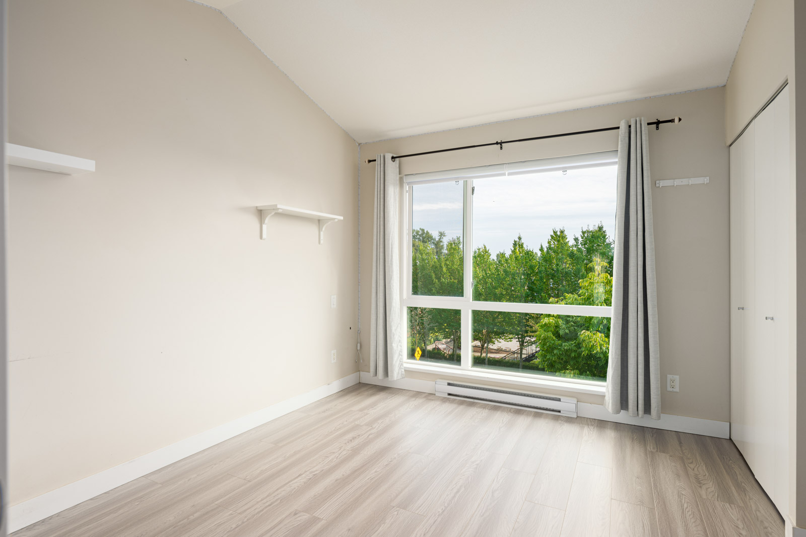 Empty room with light-colored walls and flooring, a large window with curtains overlooking green trees, a shelf on the left wall, and a closet door on the right.