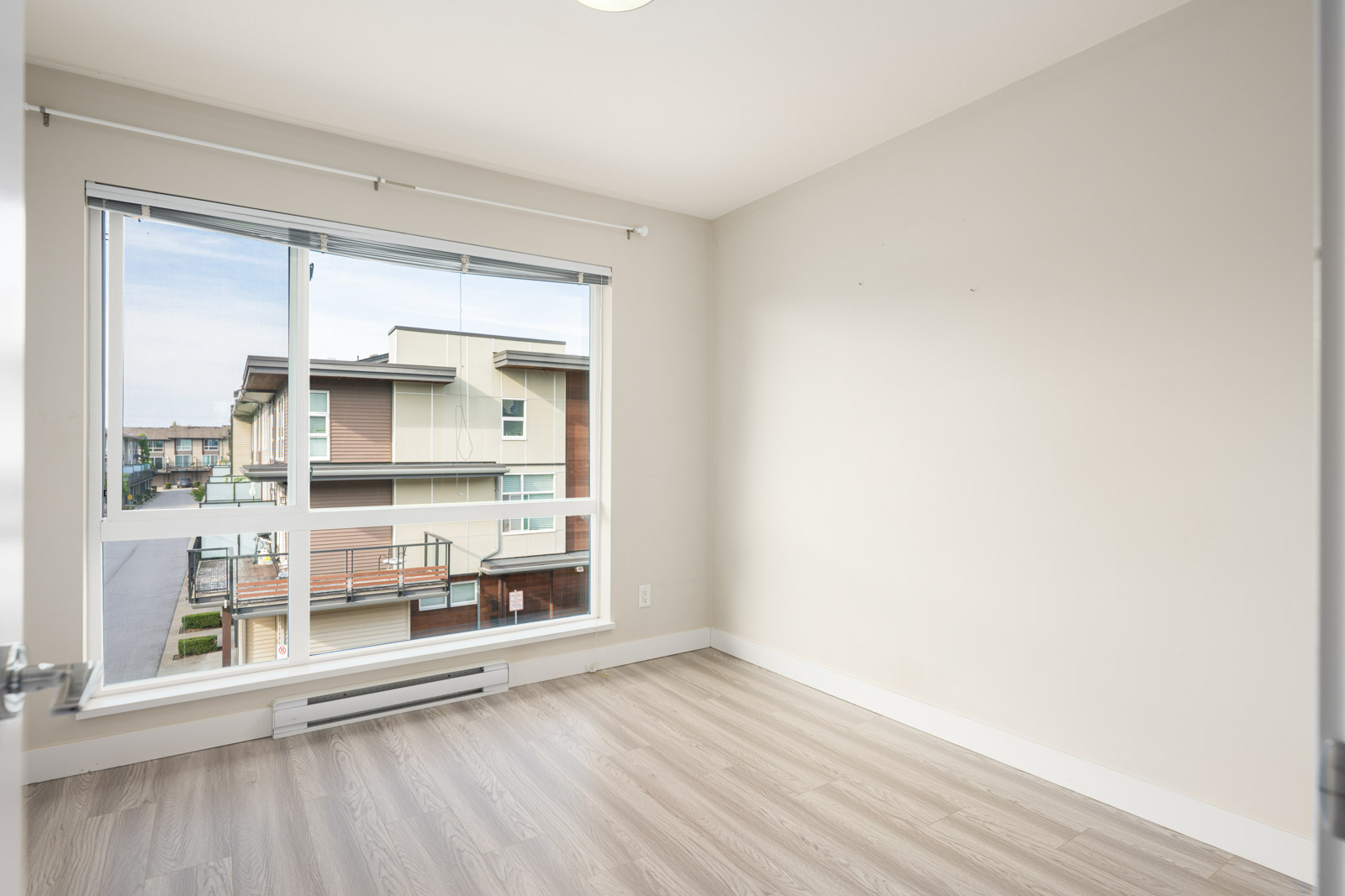 Empty room with light-colored walls and wood flooring, large window with blinds, view of neighboring modern buildings outside.
