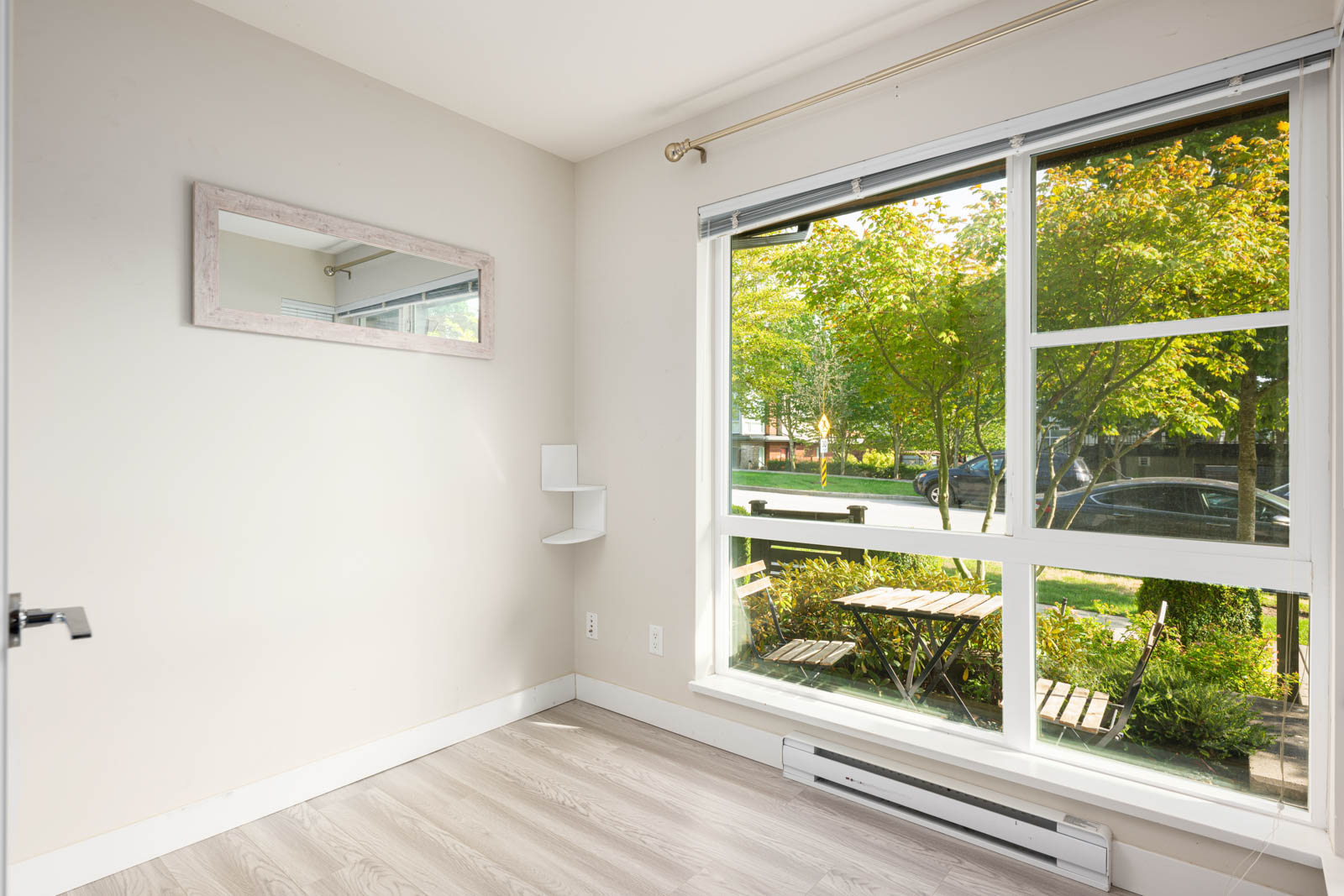 Small, empty room with light-colored walls and flooring, large window facing a patio with outdoor table and chairs, and a wall mirror reflecting the space.
