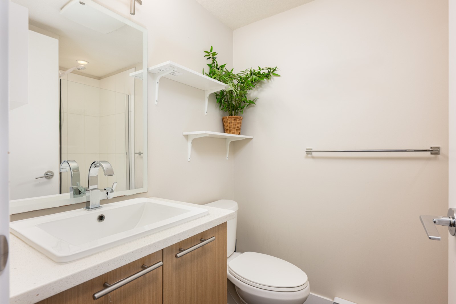 Modern bathroom with a white sink, wooden vanity, toilet, towel rack, wall shelves, and a potted plant on the upper shelf.