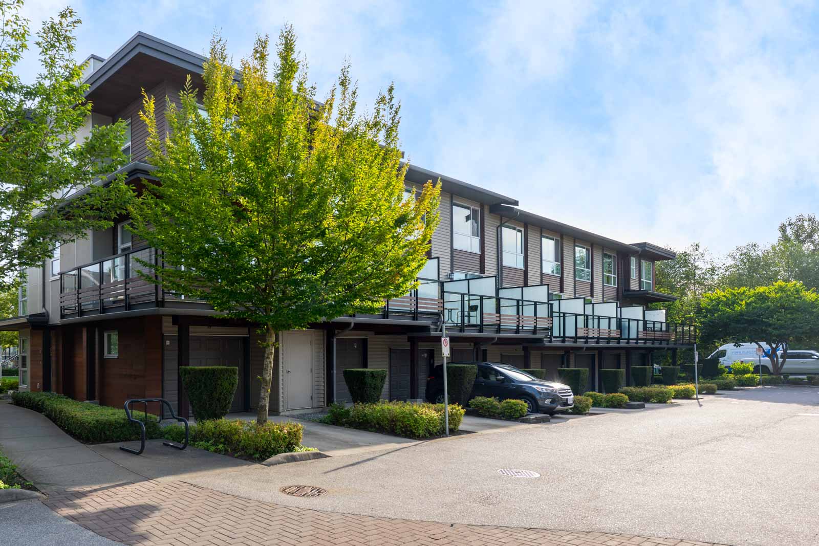 Modern two-story apartment building with large windows and balconies, surrounded by greenery and a parking area with parked cars.