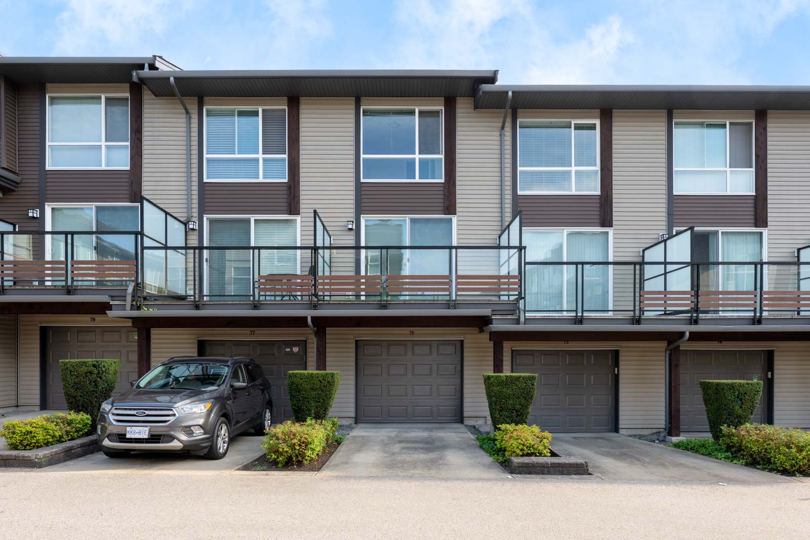 A row of modern townhouses with balconies and garages; one SUV is parked in a driveway, and manicured bushes line the entrance.