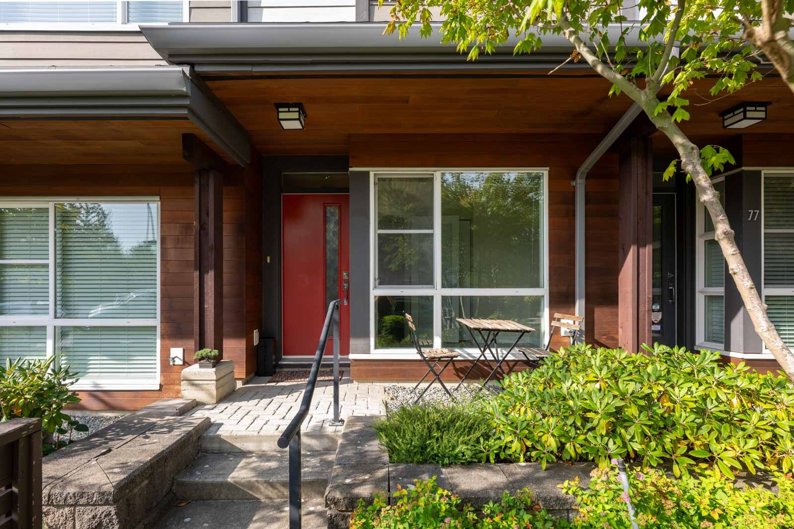 A townhouse entryway with a red door, large windows, a small patio table with two chairs, and surrounding greenery.