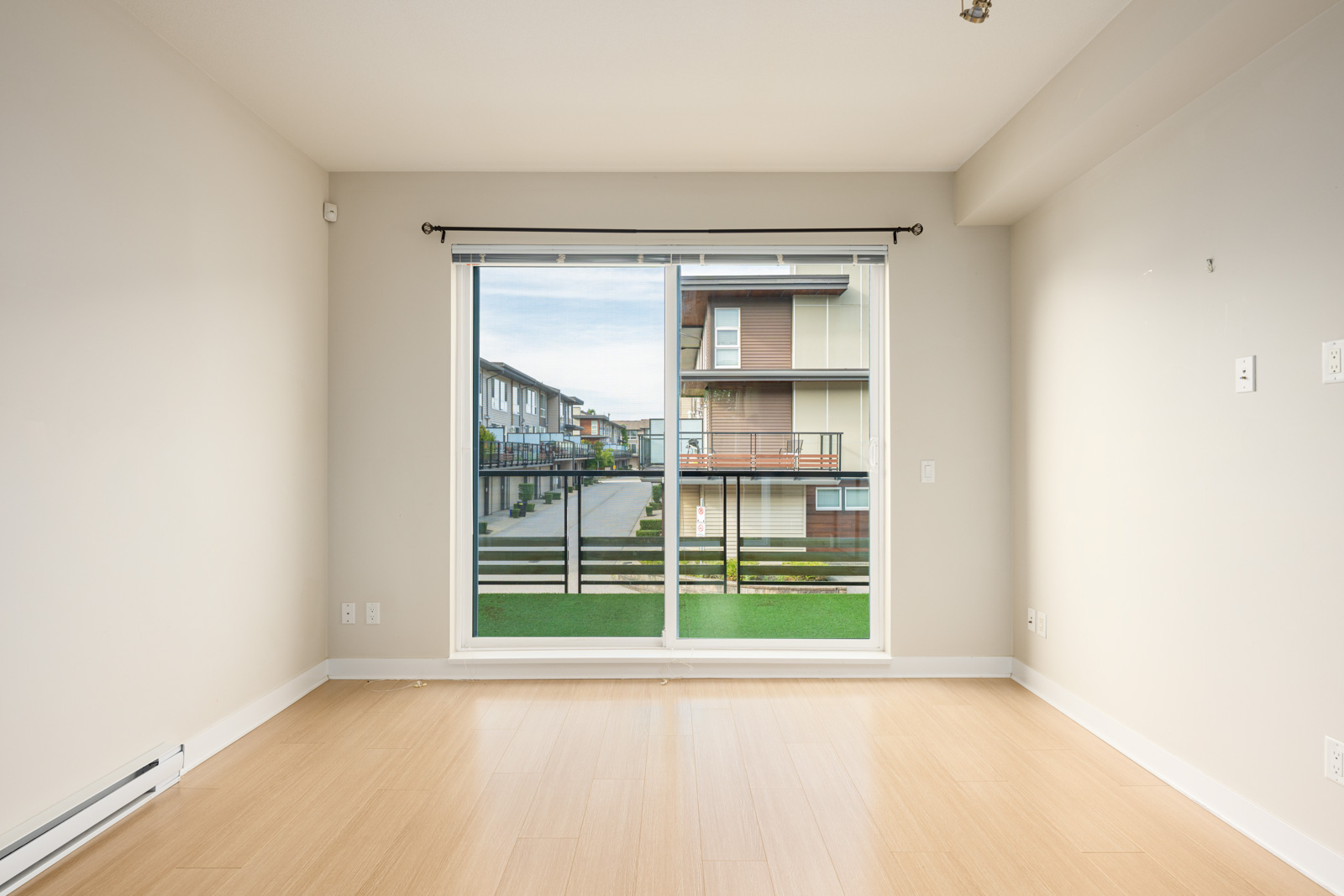 Empty room with light wood flooring, white walls, and a large sliding glass door leading to a balcony with a view of neighboring modern apartment buildings.
