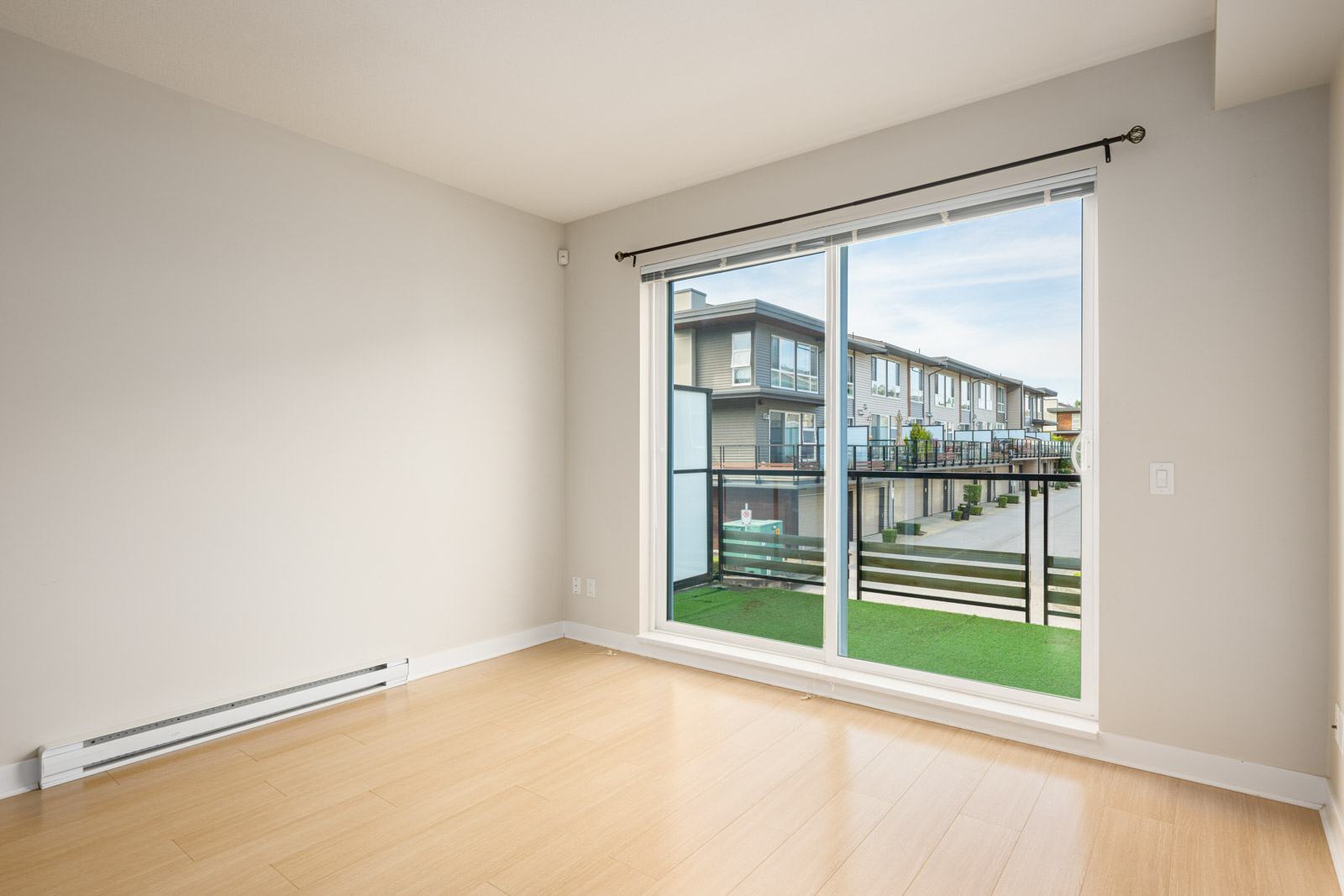 Empty room with light wood flooring, white walls, and a large sliding glass door leading to a small balcony overlooking a residential complex.