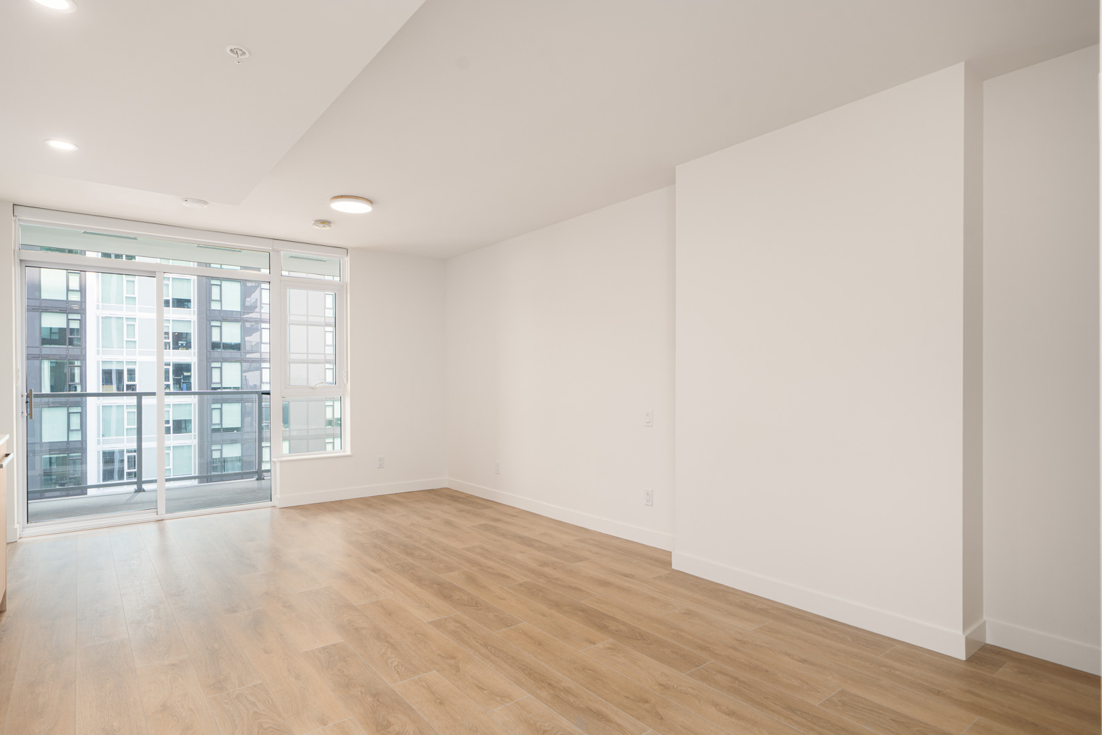 Empty modern apartment room with light wood flooring, white walls, large windows, and a glass door leading to a balcony with a view of another building.