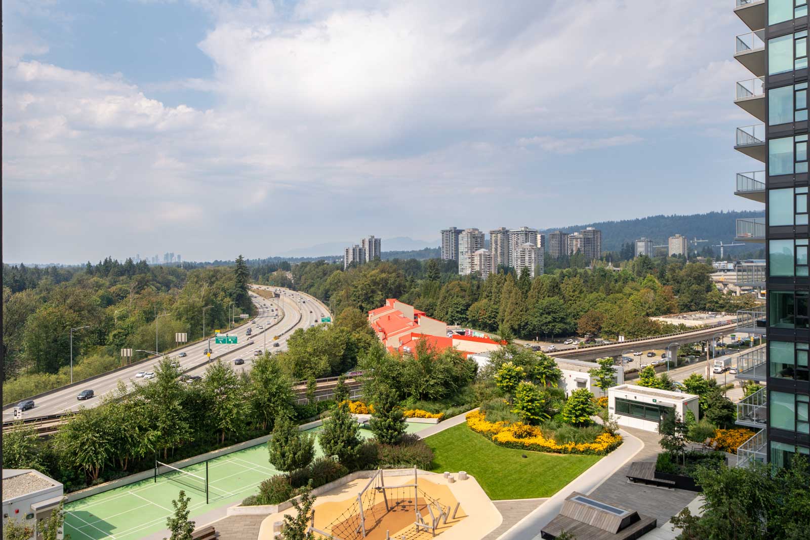 High-angle view of a cityscape with a highway, high-rise buildings, trees, a green tennis court, and a playground in the foreground under a partly cloudy sky.