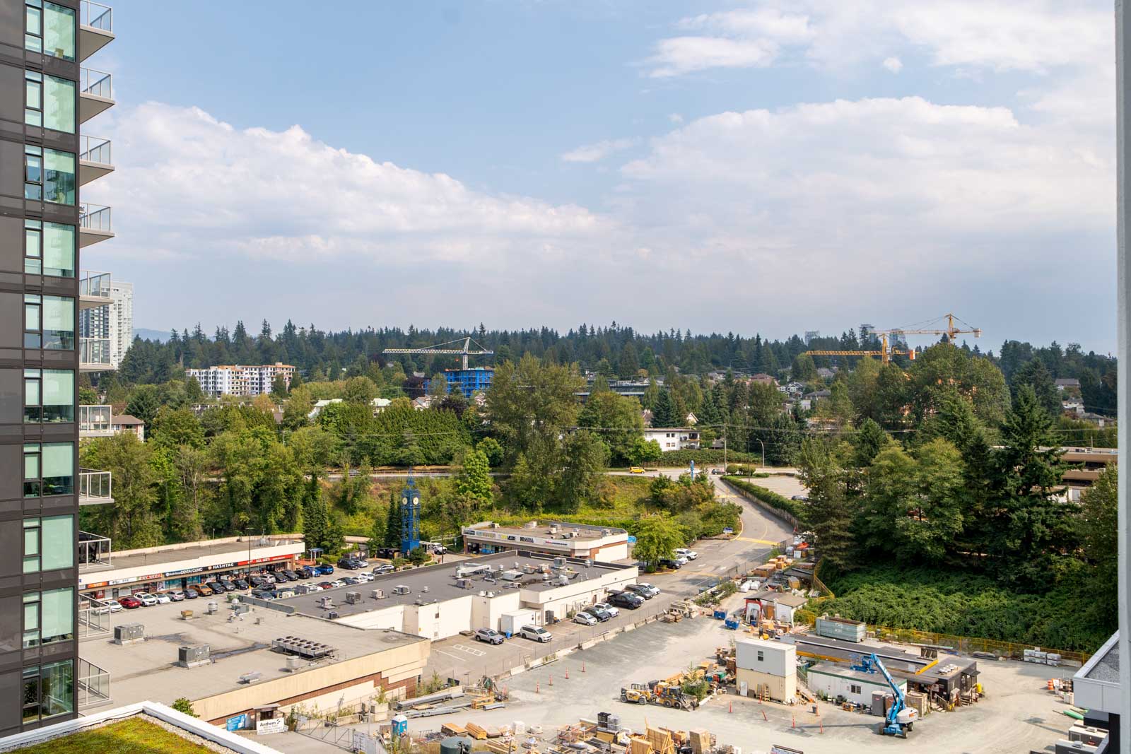 View of a construction site with equipment and materials, surrounded by trees, buildings, and a parking lot under a partly cloudy sky.