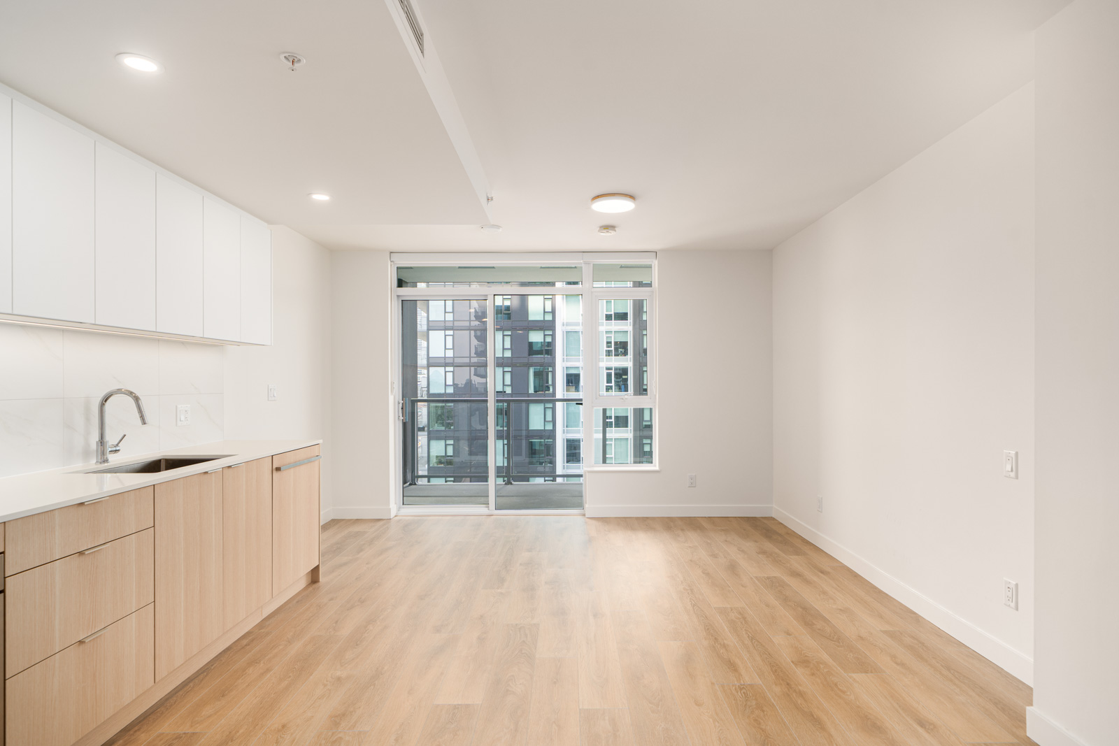 Modern, unfurnished apartment with light wood flooring, white cabinets, built-in sink, and large windowed door leading to a balcony overlooking another building.