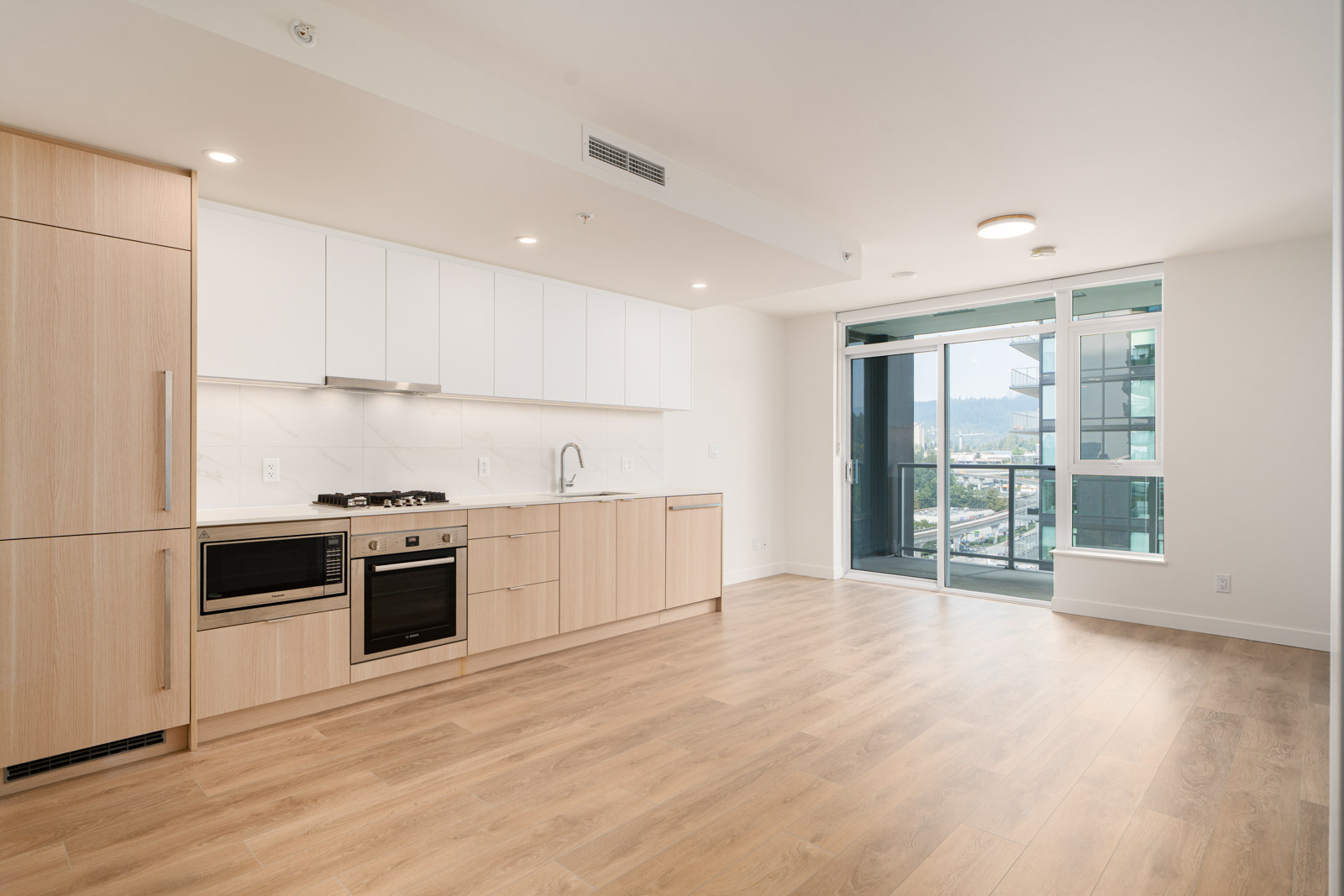 Modern, unfurnished apartment kitchen with light wood cabinets, built-in appliances, and large windows leading to a balcony with city views.