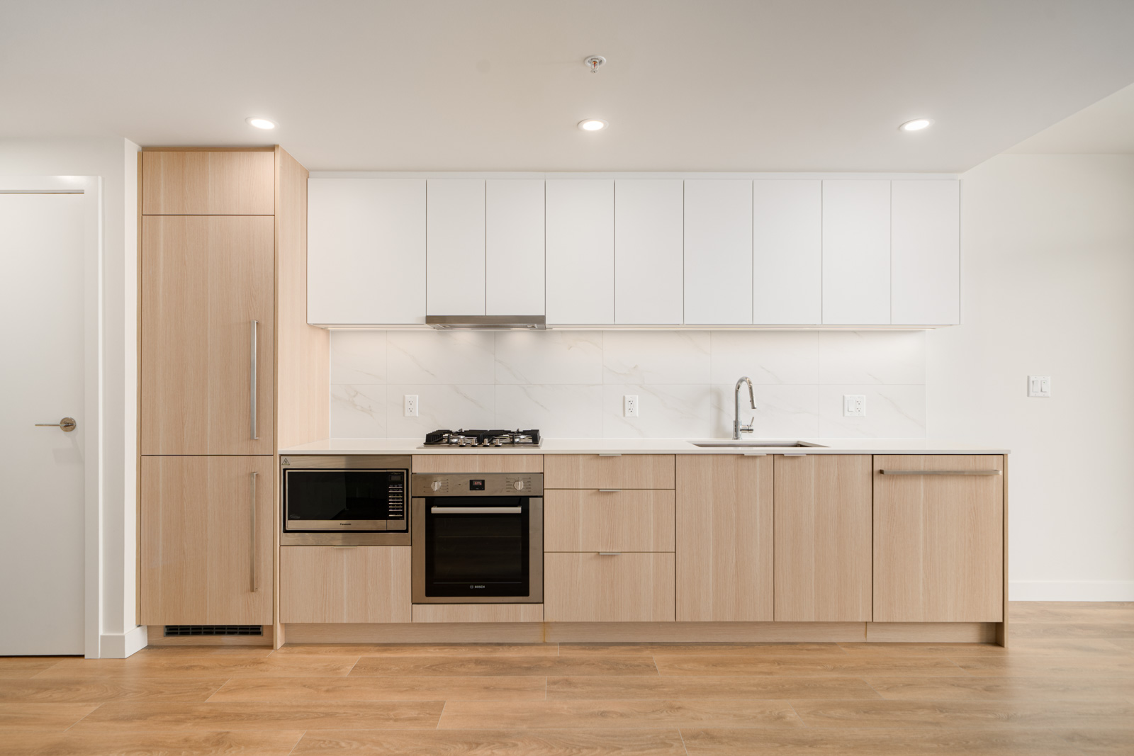 Modern kitchen with light wood cabinets, white upper cabinets, built-in appliances, gas cooktop, oven, microwave, sink, and under-cabinet lighting.
