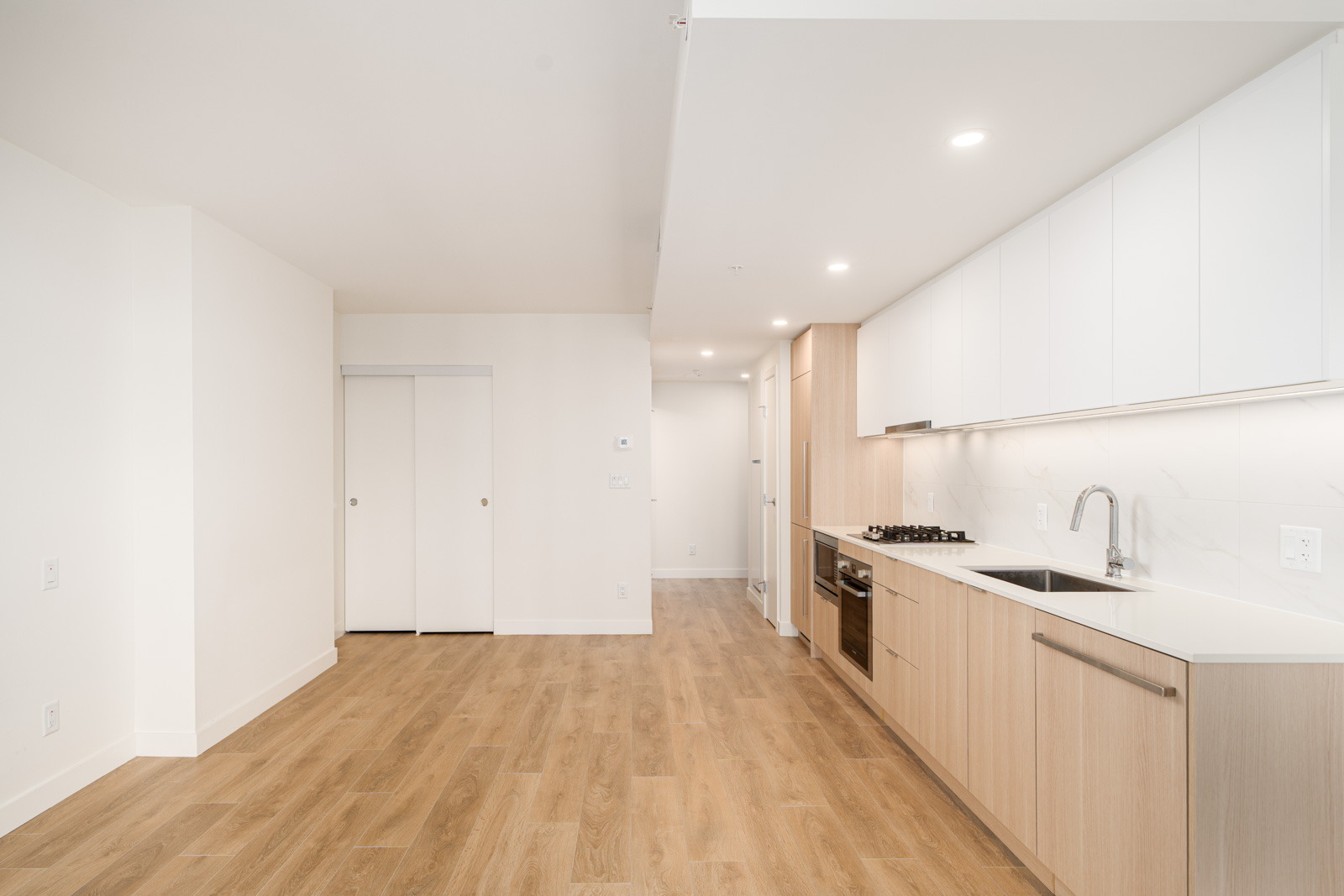 Modern kitchen with wood floors, light wood and white cabinetry, built-in stove, and sink; open area leads to a hallway and a sliding door closet.