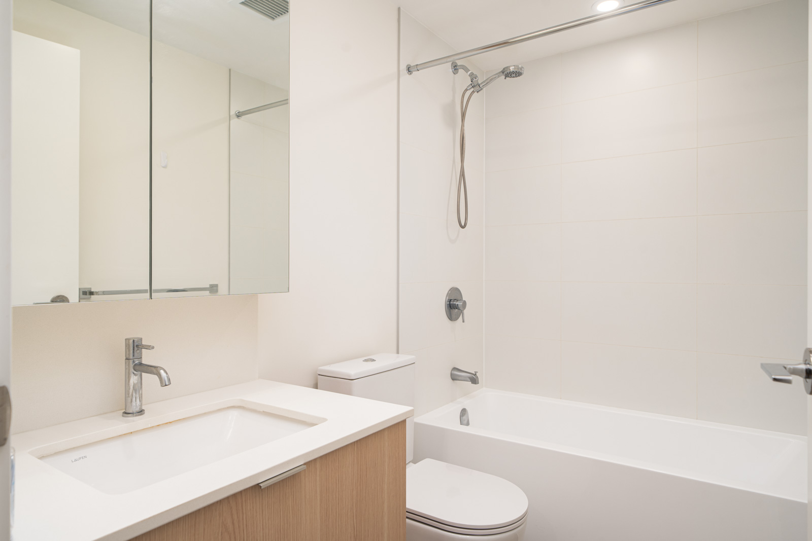 Modern bathroom with a white sink, wooden vanity, wall mirror, toilet, and a bathtub with a showerhead under recessed lighting.