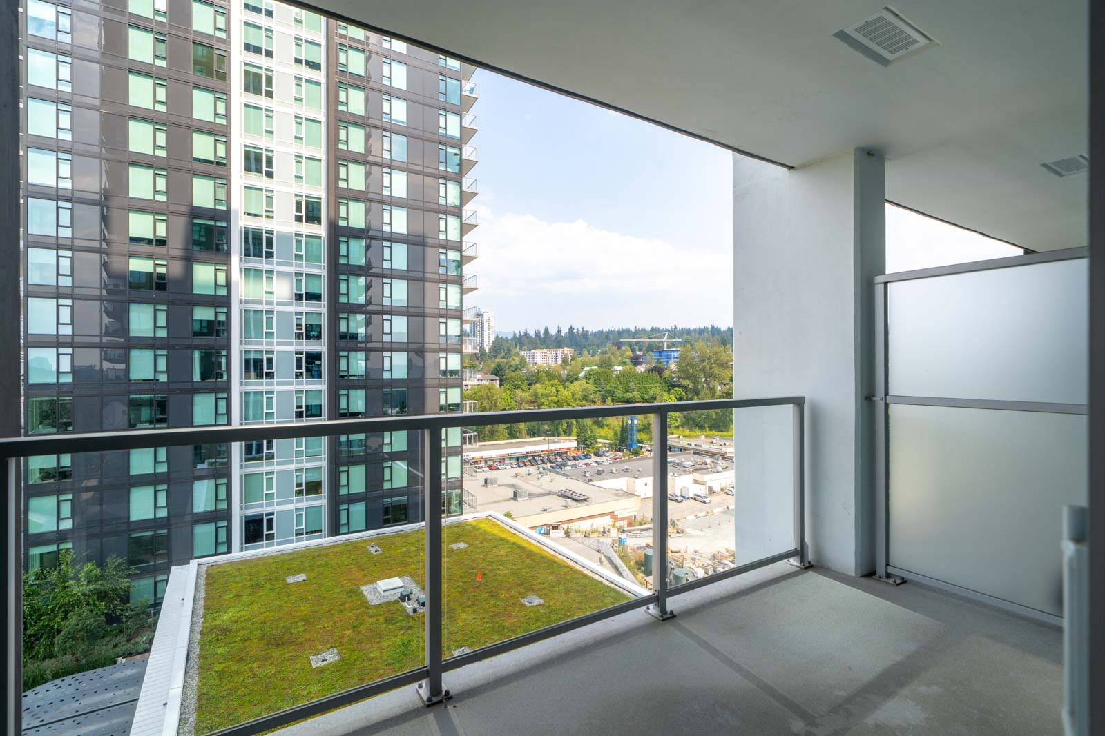 A high-rise apartment balcony with a glass railing overlooks a green roof, nearby buildings, and a cityscape with trees in the background.