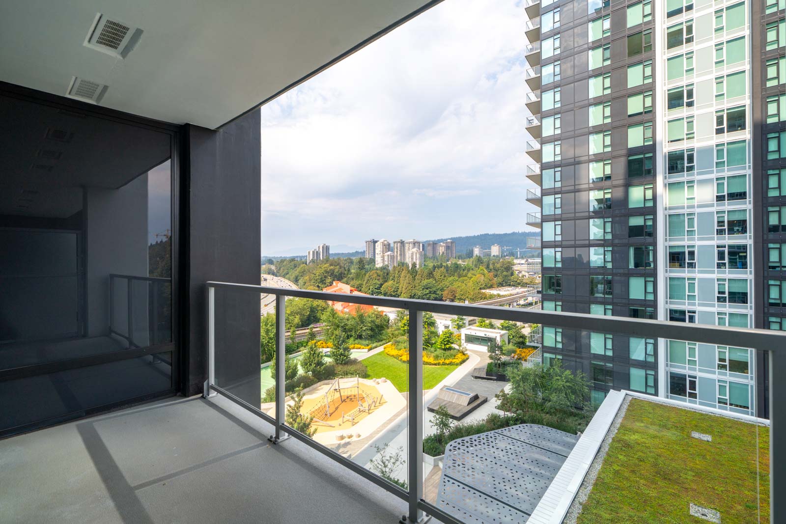 View from a high-rise balcony showing a nearby apartment building, a green roof, playground, and cityscape with trees and distant buildings under a partly cloudy sky.