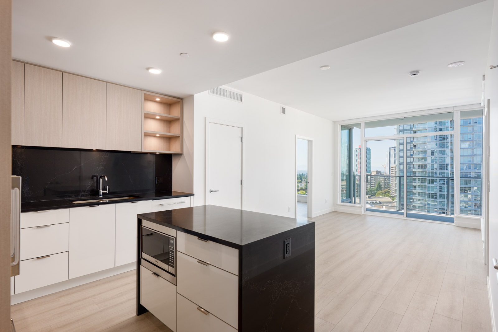 Modern, unfurnished open-plan apartment with light wood floors, a kitchen featuring a black island, and large windows offering city views.