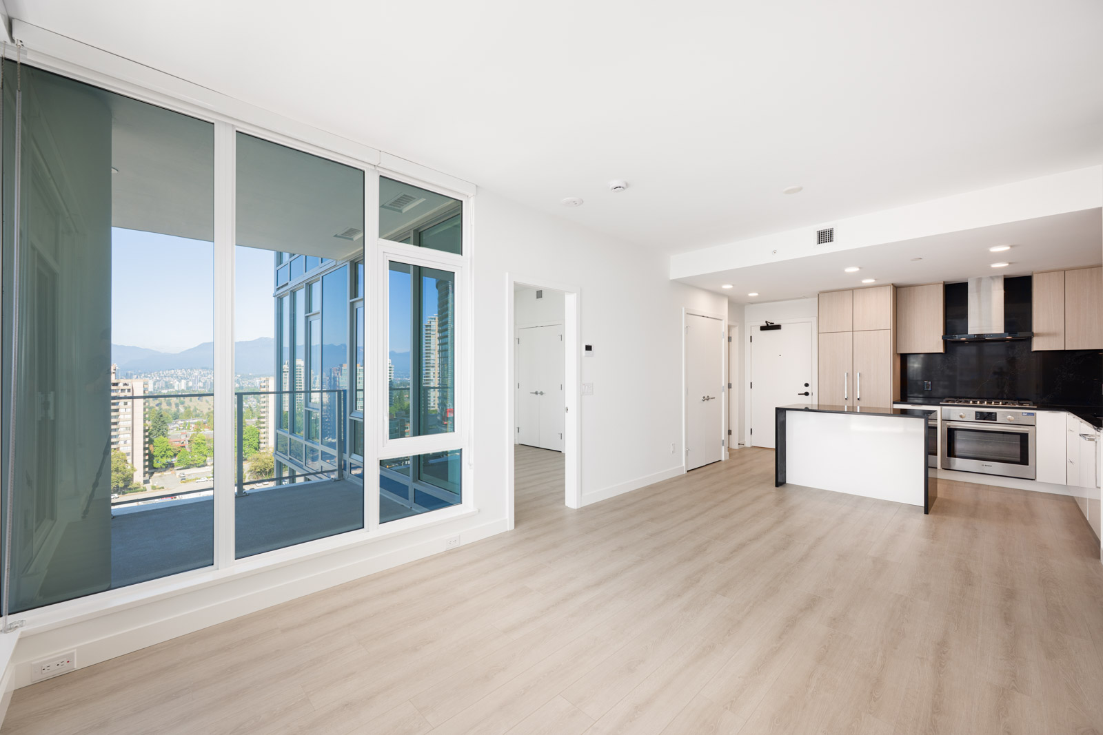 Modern, unfurnished apartment with light wood flooring, open-plan kitchen, large windows, and a balcony overlooking a cityscape with mountains in the background.