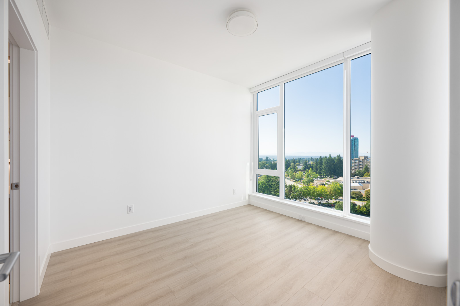 Empty, bright room with light wood flooring, white walls, and large floor-to-ceiling windows overlooking a cityscape and greenery on a clear day.