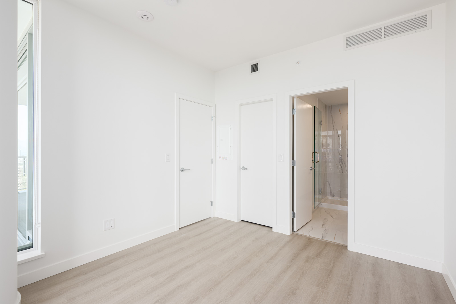 A bright, empty room with light wood floors, white walls, two closed doors, and an open door leading to a bathroom with a glass shower.