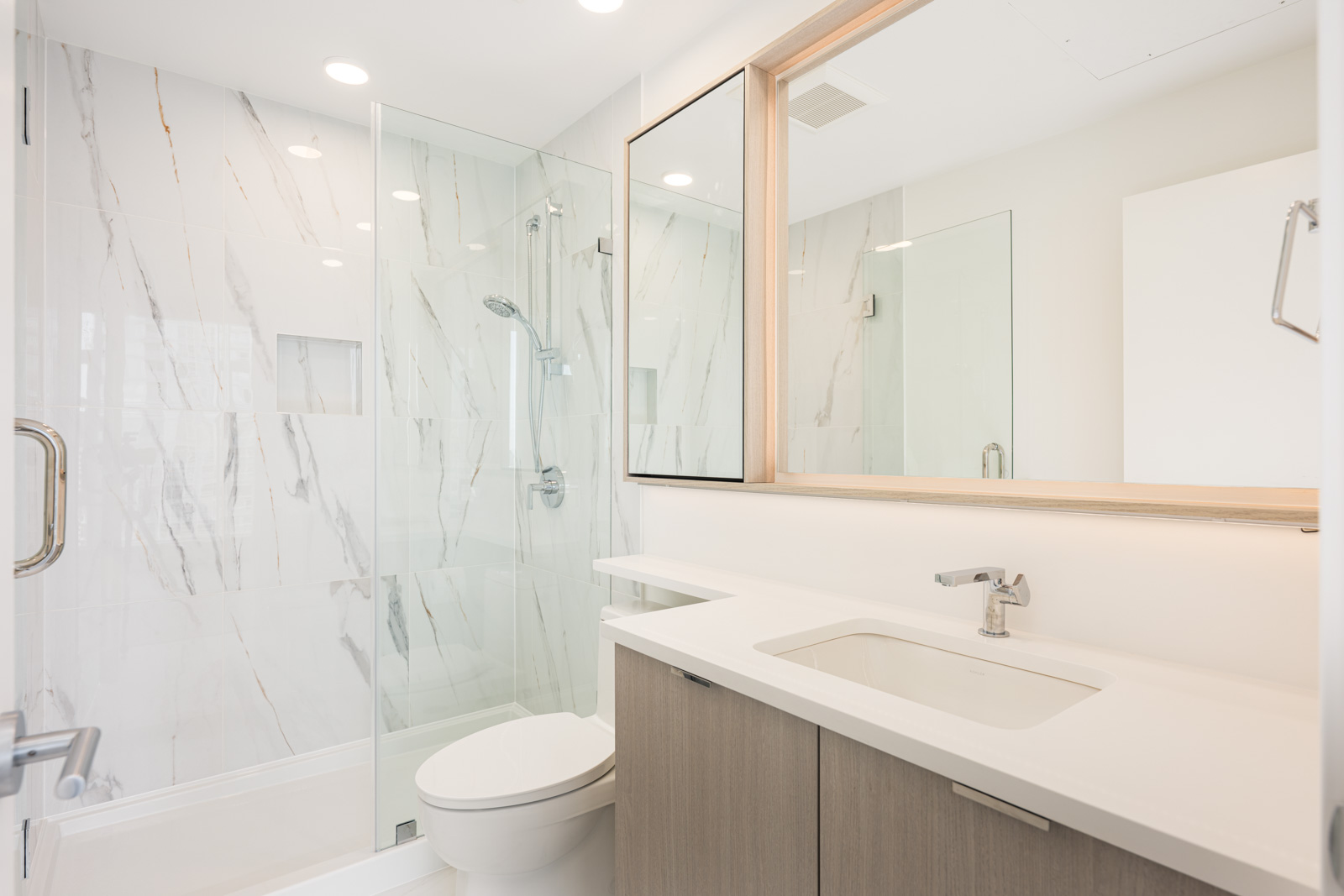 Modern bathroom with a glass-enclosed shower, marble-patterned walls, a toilet, and a vanity with a sink and large mirror under bright recessed lighting.