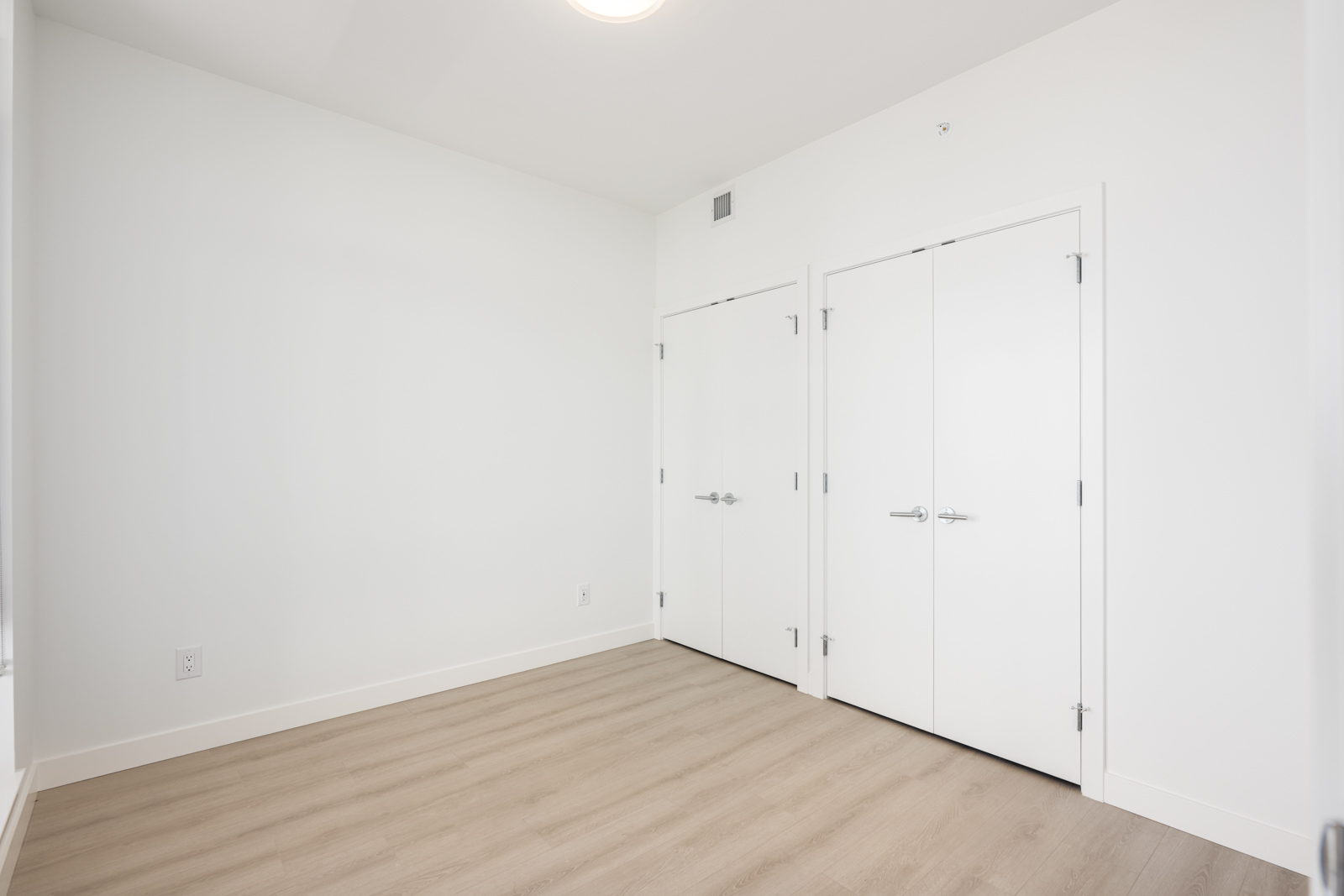 Empty room with light-colored wood flooring, white walls, and two closed white double doors. The space is well-lit by natural light from an unseen window.