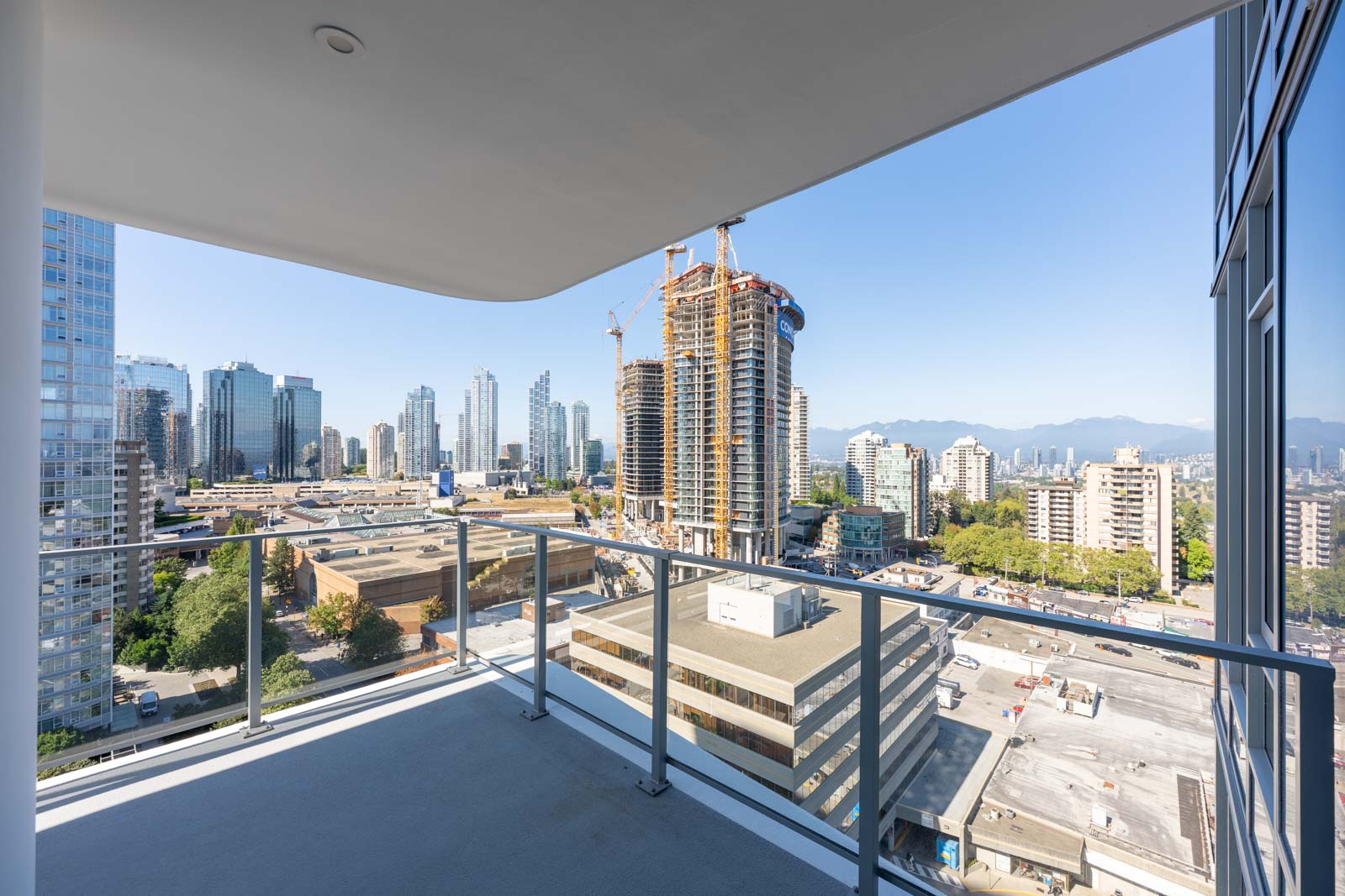 View from a high-rise balcony overlooking a cityscape with construction cranes, tall buildings, and mountains in the distance under a clear blue sky.