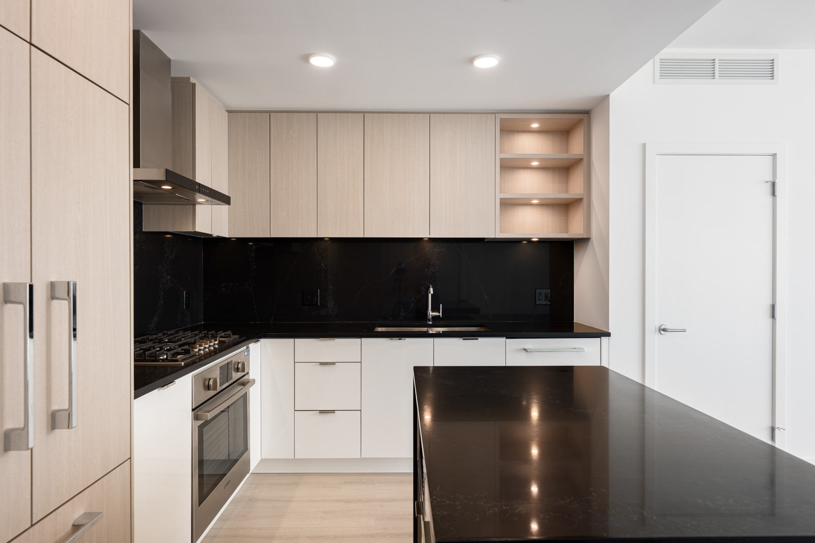 Modern kitchen with light wood cabinets, black countertops, built-in oven, gas stove, sink, and open shelves, all under recessed ceiling lights.