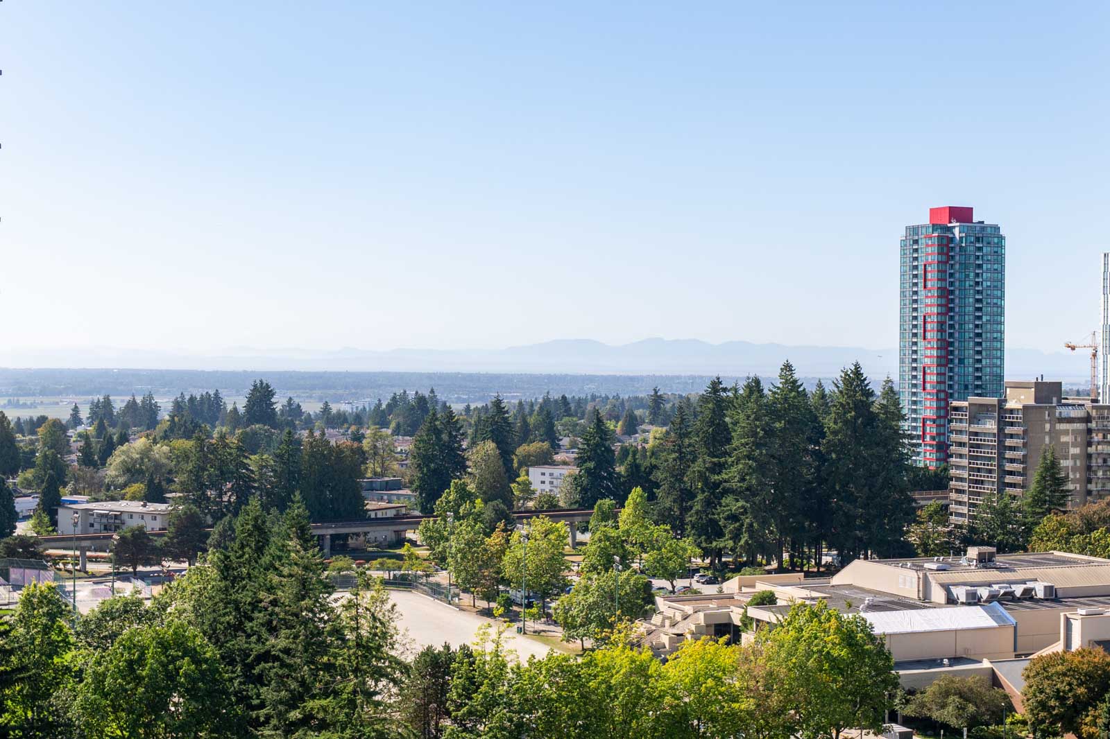 A cityscape with mid-rise buildings, a tall blue and red high-rise, trees, and distant mountains under a clear sky.