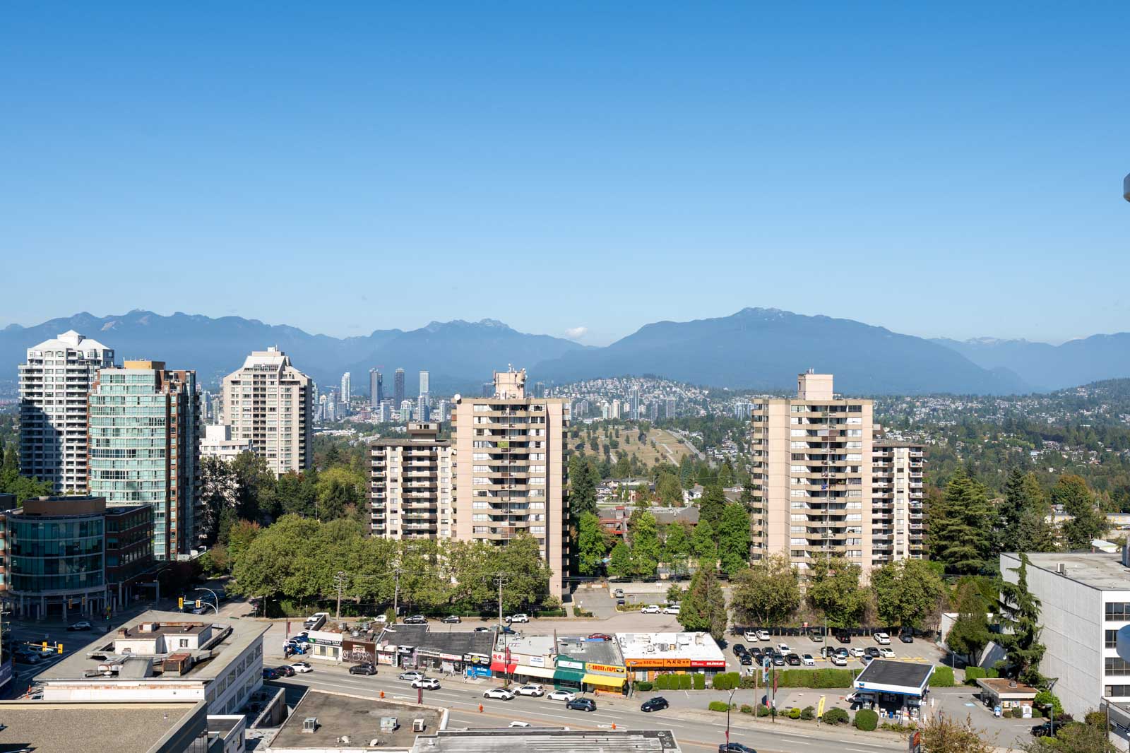 A cityscape showing mid-rise apartment buildings, roads, and trees, with mountains visible in the distant background under a clear blue sky.