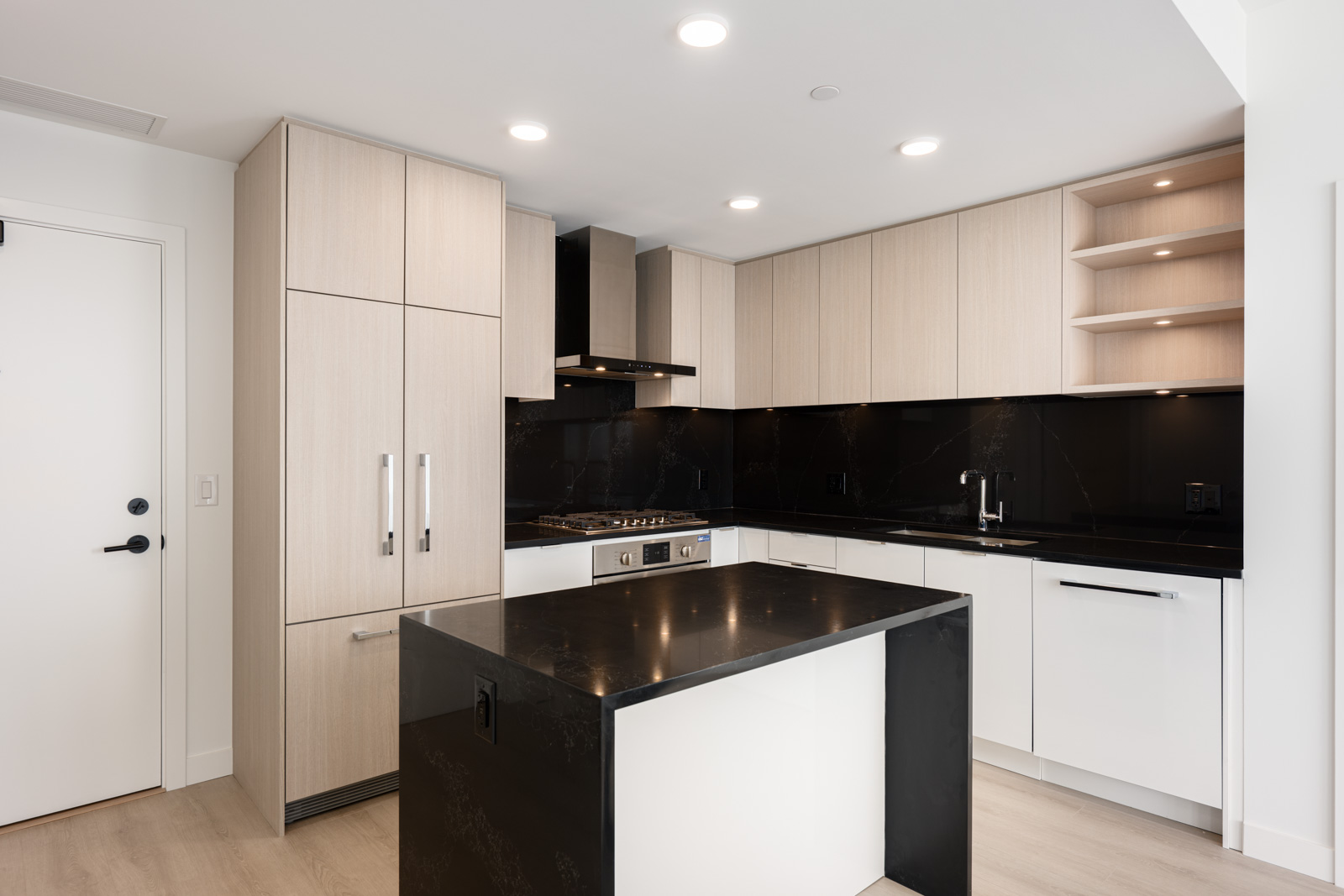 Modern kitchen with light wood cabinets, black countertops and backsplash, built-in oven, induction cooktop, island, and white walls. Recessed ceiling lights provide illumination.