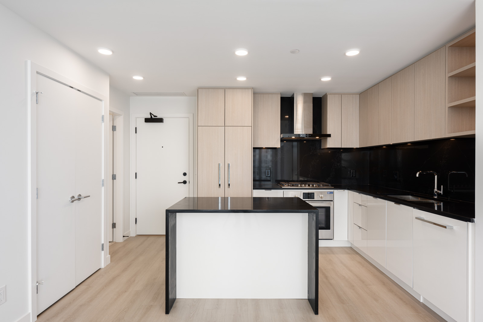 Modern kitchen with light wood cabinets, black backsplash, white island, built-in appliances, and recessed ceiling lights.