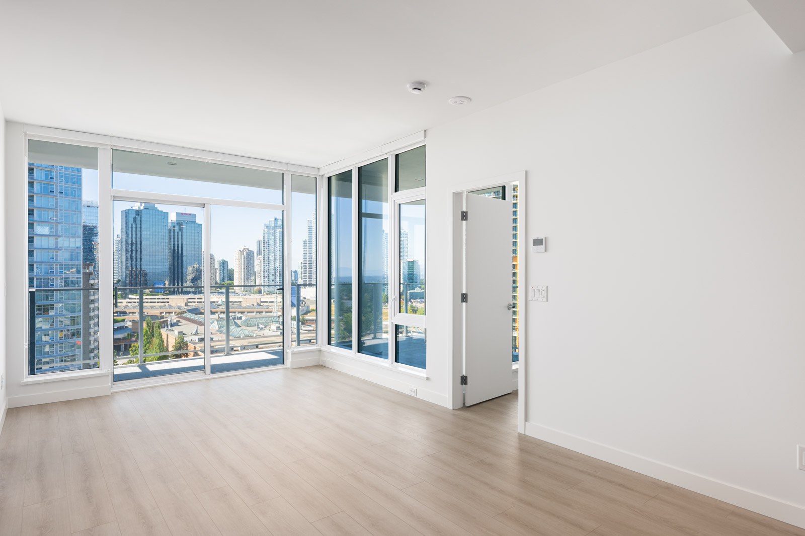 Bright, empty modern apartment room with light wood floors, white walls, large windows, and a city skyline view.