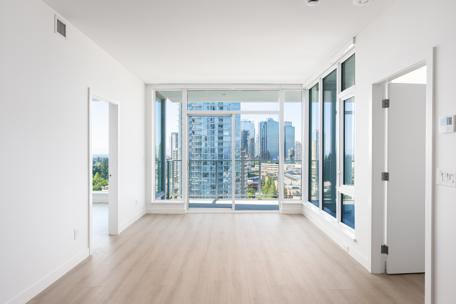 Empty modern apartment with light wood floors, white walls, large windows, and a balcony overlooking a city skyline with tall buildings.