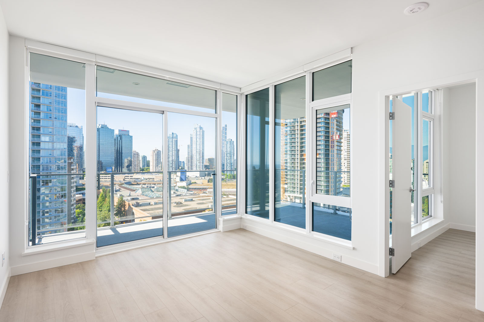 Modern empty apartment room with large floor-to-ceiling windows, light wood flooring, and a city skyline view from the balcony.