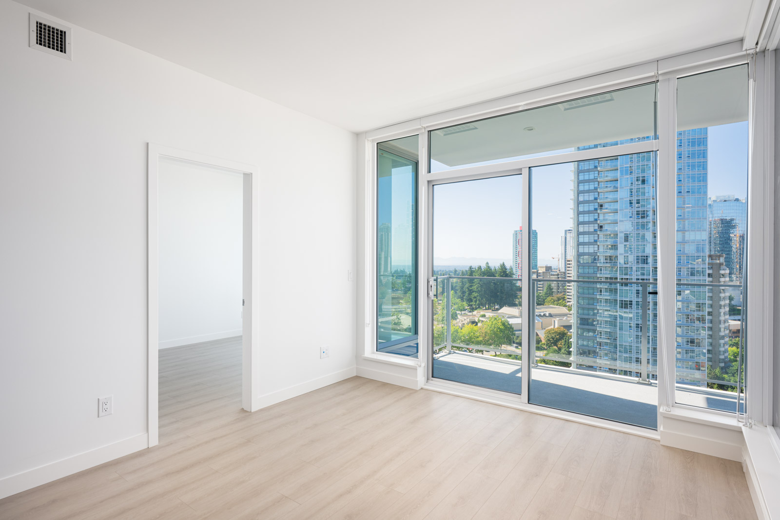 Bright, empty modern apartment room with light wood floors, white walls, and large floor-to-ceiling windows leading to a balcony overlooking city buildings.