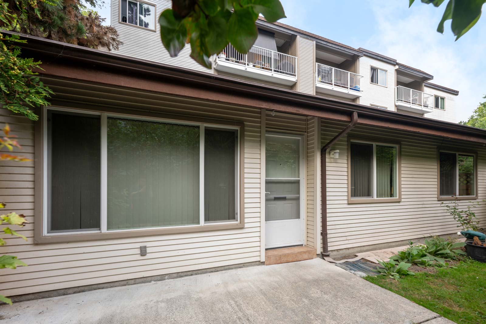 A beige two-story townhouse with large windows, a white door, and a concrete walkway, surrounded by some greenery and plants.